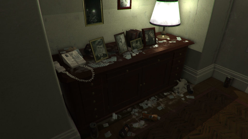 In the screenshot taken from the P.T. Game we see a messy desk made out of dark wood in front of us. On it is a telephhone with a cord that is not hung up, some black and white photos of people in frames and many drugs all around it. On the floor are used beverages and a white teddy bear.