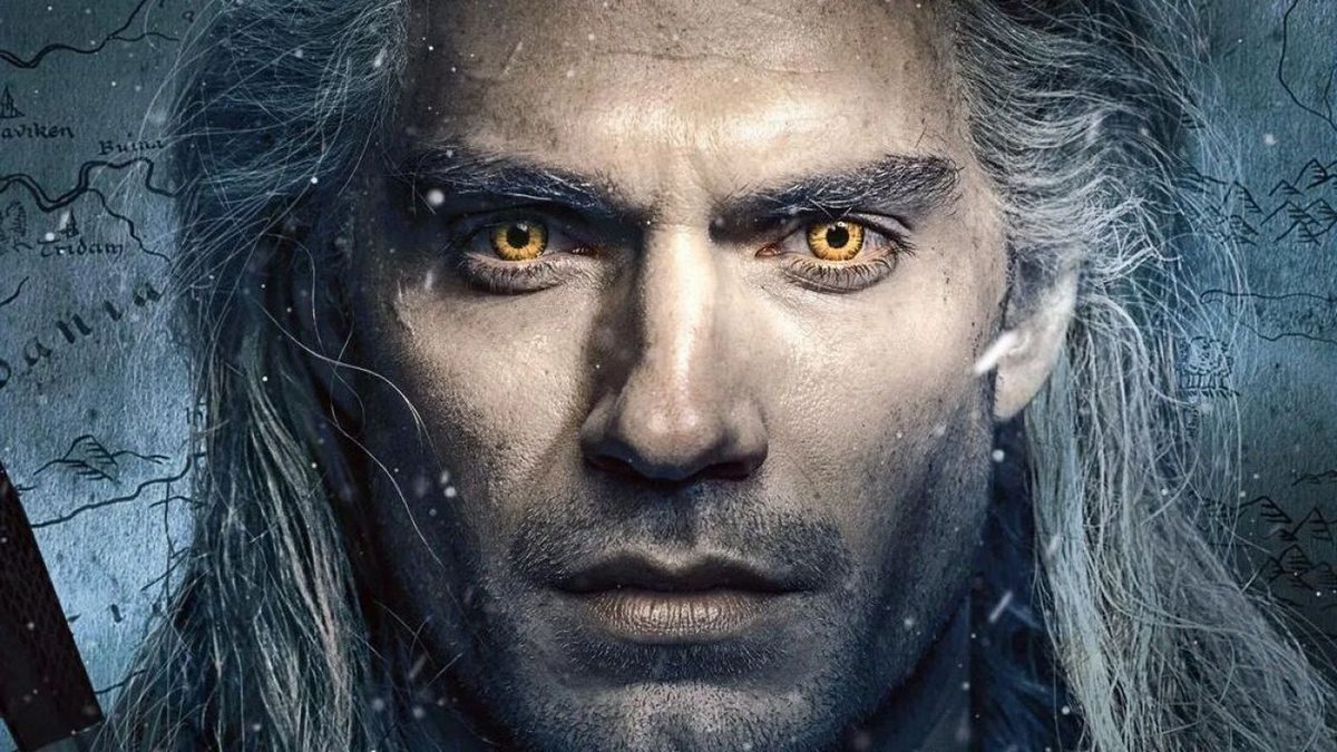 In this image, we see the face of the protagonist Henry Cavill in a close-up from the front, so that almost the entire frame is taken up by it. The actor is part of the cast of The Witcher season 3. He has a shaved face, and long white open hair and his eyes look at us with an orange glowing iris. In the background, we can see a drawn black-and-white map with mountain symbols and strange writing. His face is illuminated with a warmer light, but the background appears dark silver.