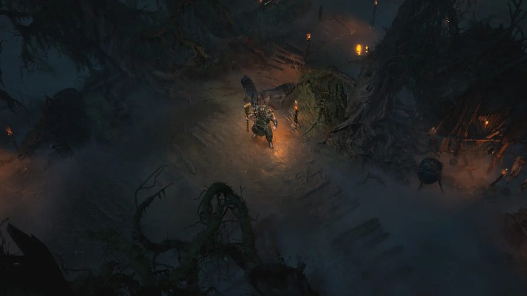 This screenshot from Diablo 4 shows the Hawezar region at night. In the center of the image is a playable character in a wooded, village setting, which is sporadically lit by torches and also illuminates him somewhat. Around the player we see tall trees with large roots and twisted branches, which are also visible in the foreground in the crop. This screenshot is not part of the leak, but it is a screenshot published by Blizzard.
