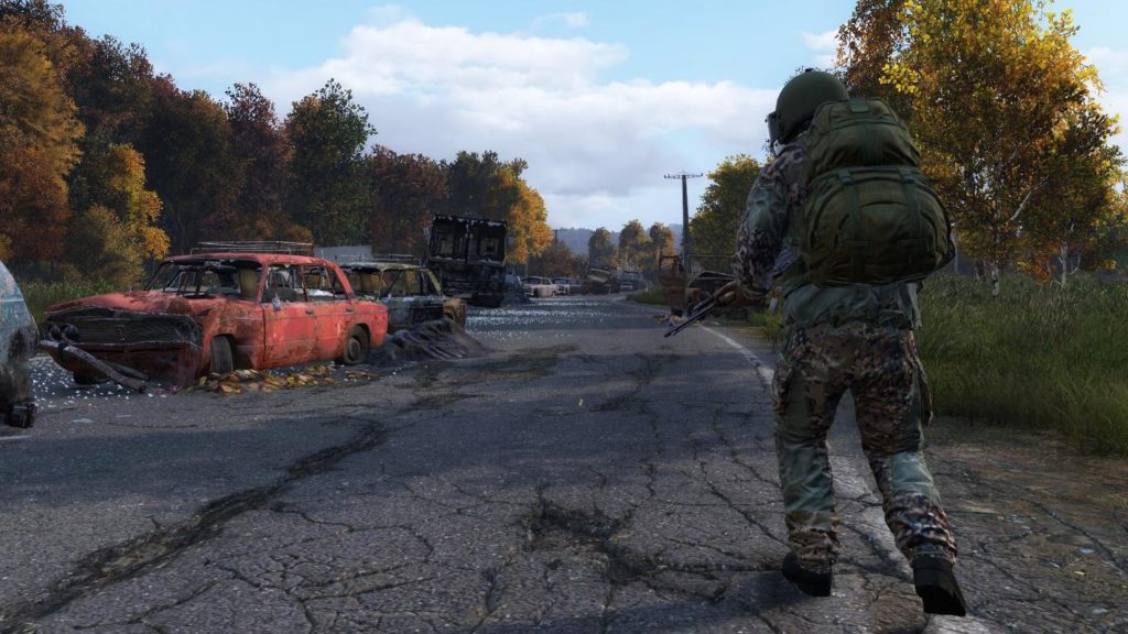 In this image, which is a screenshot from the game DayZ, a player armed with a Kalashnikov is walking straight down an abandoned street in daylight, where there are many destroyed cars. The player is shown in a slight bottom view from behind in the wide shot and on the right side of the image. The road runs from the foreground of the picture into the background. The dense forest is shown next to the road on both sides. The wrecked cars are seen staggered on the left side of the road, pointing in the direction of the viewer.