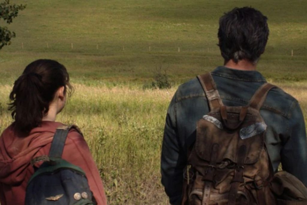 The main characters Ellie and Joel are standing on a meadow in the TV show The Last of Us on HBO. We look at them from behind and see the wide meadow over their shoulder. The green leaves of a tree can be seen in the upper left corner and a drony dark shrub in the center of the image. The meadow in front of them slopes slightly and rises again to a hill in the background.