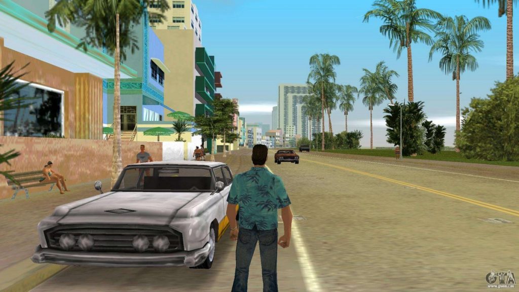 What does the GTA 6 map look like and when does GTA 6 come out? In this picture, we can see a part of GTA Vice City. We see the player in third-person view centered in the bottom half of the image, standing on a street in daylight next to a parked white car. He wears a turquoise short-sleeved Hawaiian shirt and has short black hair. His gaze falls to the center of the picture, where the road also runs like a vanishing point. A red car can be seen in the center of the picture, driving toward the player. On the right side, tall palm trees are depicted in a green park with bushes and shrubs. On the left side are several different colored buildings with balconies and windows. In front of the houses are walking people on a sidewalk and a sitting woman on a bench in the foreground of the picture. In the distance, skyscrapers and other buildings rise into the light blue sky. The GTA 6 map (leak?) should be based on this area.