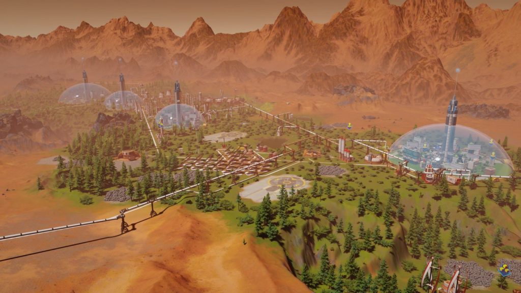 The picture shows a clip from the game Surviving Mars. In the long shot, you can see several colonies on the planet Mars, which are enclosed under huge oxygen-giving domes. Between the colonies run various pipelines and a wide stretching green landscape with green fir trees is shown. An orange dusty mountain range can be recognized in the background.