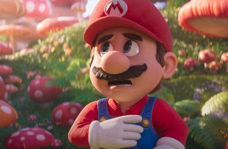 The plumber from the Mushroom Kingdom is shown in the center of the picture in the close-up with his red shirt, red cap, blue dungarees, and a brown mustache. He is standing in the middle of the day on a hilly meadow, which is covered with various large red toadstools, which we see out of focus in the background. Worried, he looks upward to the left out of the picture. At the top right of the image, a blue toadstool is also shown in the crop. This is a screenshot from the Super Mario Movie film trailer, which was released in the fall of 2022.