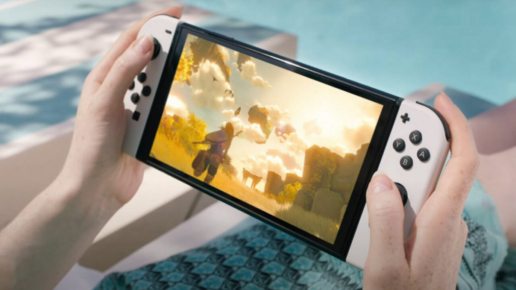 In this image, we see the Nintendo Switch in the total view from an oblique perspective with white Joy-Cons in the hands of a person lying on a lounger outdoors. Behind the Switch, the legs of the person playing can be seen wrapped in a turquoise towel and running out of the picture to the right. The towel contrasts with the white lounger we see in the crop. Further in the background, a turquoise floor is shown. The person is playing a Zelda title on the Switch, and we see the protagonist Link in third-person view at the bottom left of the screen with blond hair and a bow in his hand, running through a light-flooded yellow valley. 