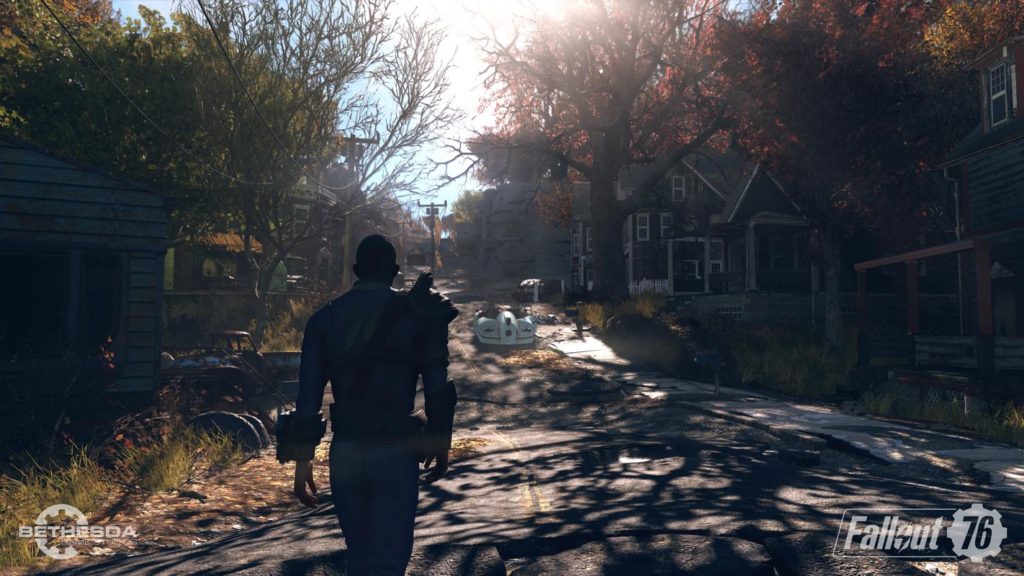 We see a screenshot from Fallout 76, where the main character is shown in the third-person view in the bottom right of the image in the semi-close-up, as he walks in daylight on a street through an abandoned city. The street runs from the front straight up a bit into the background of the picture and is surrounded by wooden houses with porches and trees with red and yellow leaves. The sun shines directly at us from above and many shadows of the trees are depicted on the street.