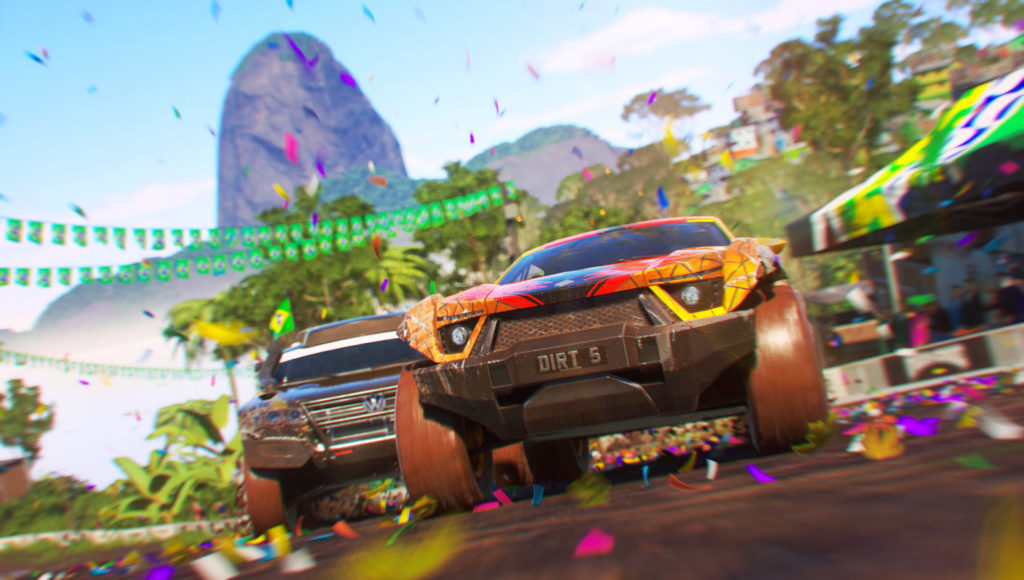 One of the best racing games for PS5 is Dirt 5, and here we can see a screenshot of two dueling drivers in an impressive frontal and bottom view perspective. The race track consists of brown sand and is surrounded by palm trees and a stage on the right side, which can be seen in the crop. Colorful confetti rains down from the sky onto the car and the track. In the distance, a large mountain rises from the forest and we see the blue sky behind it.