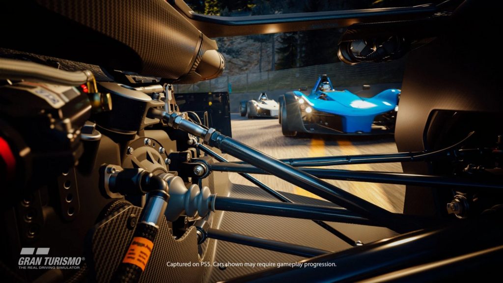 In Gran Turismo 7, one of the best PS5 racing games, you experience a very impressive racing simulation, as can be seen on this game cover: As a viewer, we take a very spectacular view here in first-person perspective. We look through the gearbox to the rear and thus see a blue racing car and a white one behind it, which are very close to our racing car. As a result, there are numerous metallic bars and mechanics in the foreground of the image. An asphalt race track with yellow markings can be seen and a large white fence behind it. 