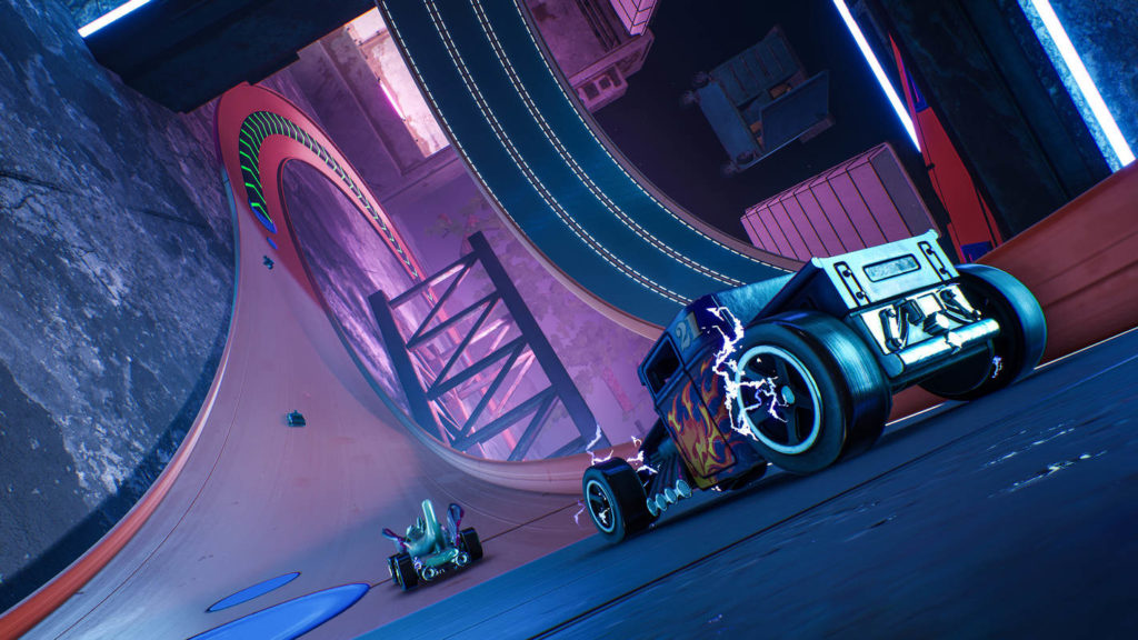 In the game Hot Wheels Unleashed, one of the best PS5 racing games, you drive toy cars. In this picture, we are in a very oblique perspective in the middle of a race track, which at this point is tilted almost 90 degrees to the left and runs in red color to the top left of the picture into the background, where it then makes a right turn. Directly in front of us, on the right half of the image, we see a blue car with a fire logo and purple flashing tires. Directly in front of it are other cars at some intervals. The course of the track is very curvy with different heights.