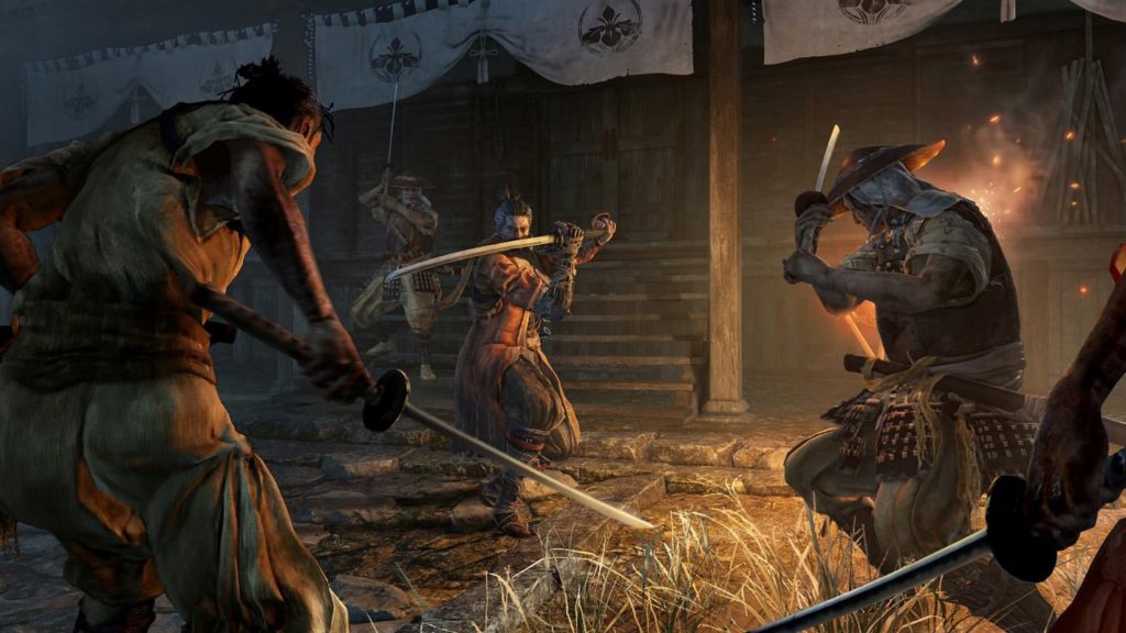 Here we can see a screenshot from the game Sekiro: Shadows Die Twice. We are looking at a dark stone street illuminated by fire from an oblique perspective. The entrance of a wooden, elongated building can be seen behind it. The scene is set in Japan. The protagonist of the picture, a shinobi named Sekiro, is shown from the front in the center of the image in a long shot with his katana sword raised as he slowly approaches two enemy characters, shown from the back on the left and right in the foreground, who also have their swords drawn. In the lower-left corner, we see a hand with a katana in the gating. 