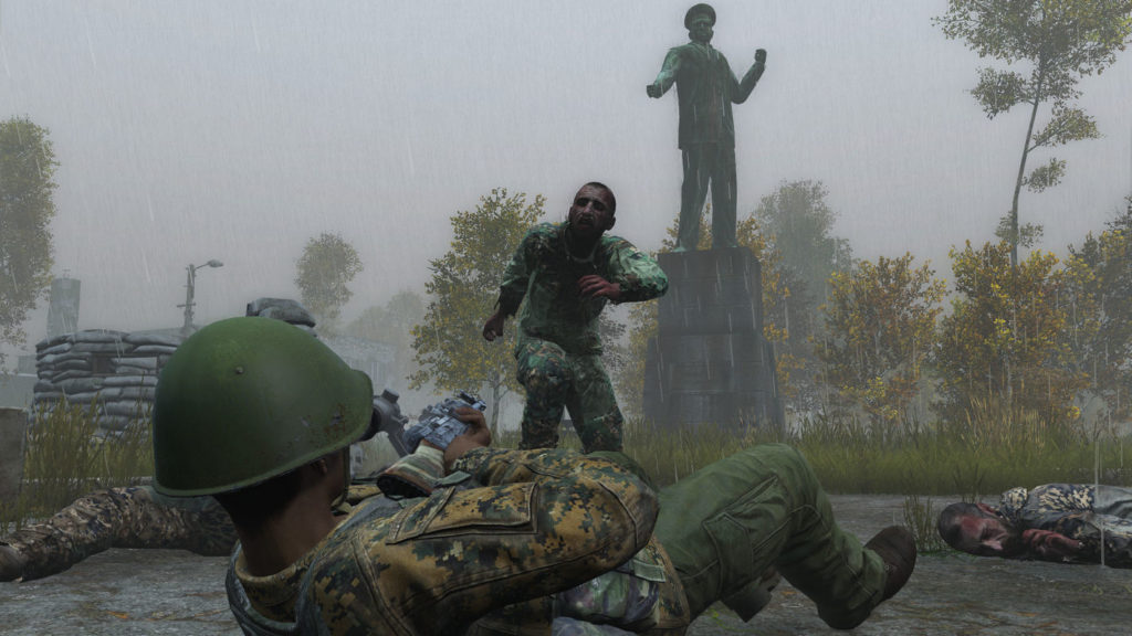 Try to fight for your life in one of the most popular survival horror games. In this screenshot, we can see the player in a third-person view in a Russian uniform and helmet, holding a Kalashnikov in his hand. He is in the middle of a gray, rainy, Russian city. In the center of the image, a zombie in a soldier's uniform is shown running toward the player who is about to shoot him with his rifle. To the left and right of him, dead zombies are already lying on the ground. In the background, you can see a bunker made of sandbags on the left, a large brass statue in the middle, and several trees around it on a lawn.