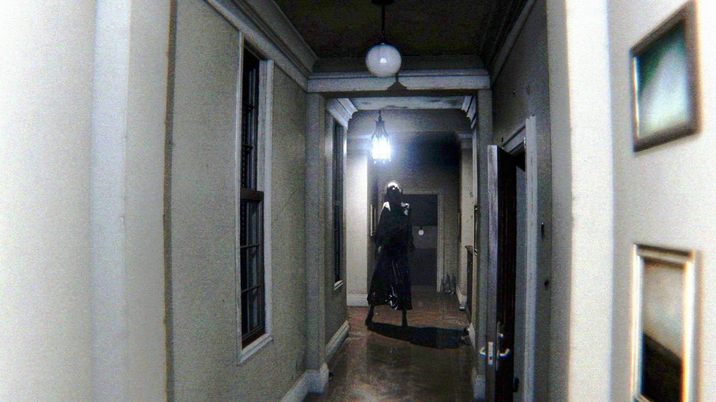 This screenshot from the original P.T. game shows a long hallway with an creepy tall person standing at the end of it, looking at us. In front on the right wall is an slightly open door, further back an eerie lamp hangs from the ceiling and there the hallway splits to the left and right. Behind it is another open door.