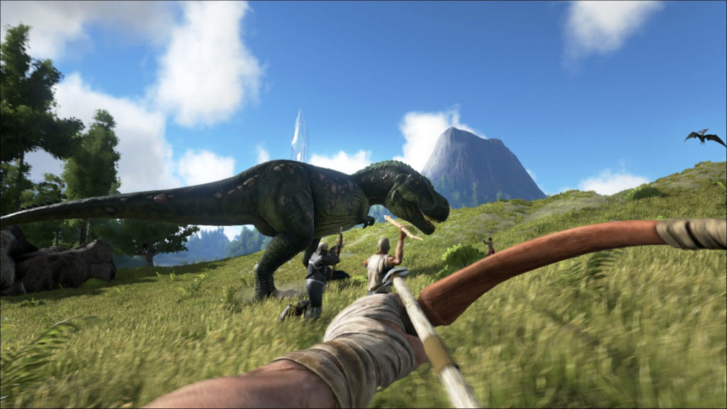 Play Ark: Survival Evolved on PlayStation 5 and hunt dinosaurs. In this screenshot, we're aiming a tense bow in first-person perspective at a T-Tex standing on a green field, which we're also standing on. The dinosaur is shown in profile in the middle ground of the image, running to the right. Directly in front of it are two other players. The one on the left is armed with a bow, and the one on the right with a spear, who are also attacking him. On the left edge of the image, a green forest can be seen in the crop, and a volcano in the distance on the right. Above it, we glimpse the bright blue sky with a few white clouds. This game is one of the highly recommended PS5 2 player games.