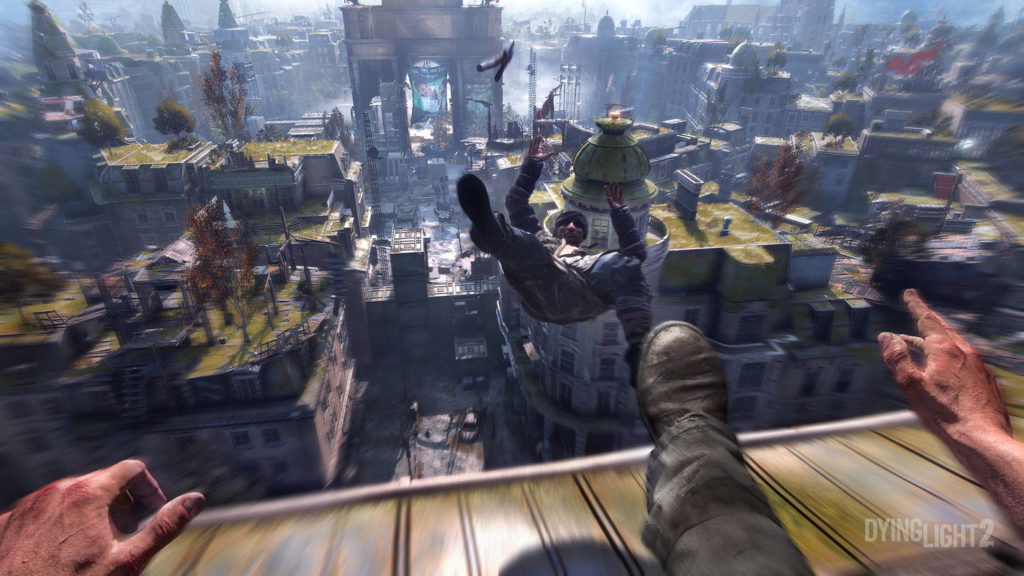 Jump across rooftops with friends and fight hordes of zombies in one of the most remarkable co-op games for PS5. Here we see a screenshot from Dying Light 2: Stay Human. The image shows our view as a player in the first-person perspective, as we are currently standing on a rooftop in a post-apocalyptic city in the middle of the day. Our right foot sticks out into the picture because we are kicking an enemy zombie off the roof, which can be seen in the center of the picture still in the air with its arms and legs outstretched. In the background, we see a devastated city with its numerous houses and roofs in a slight motion blur.