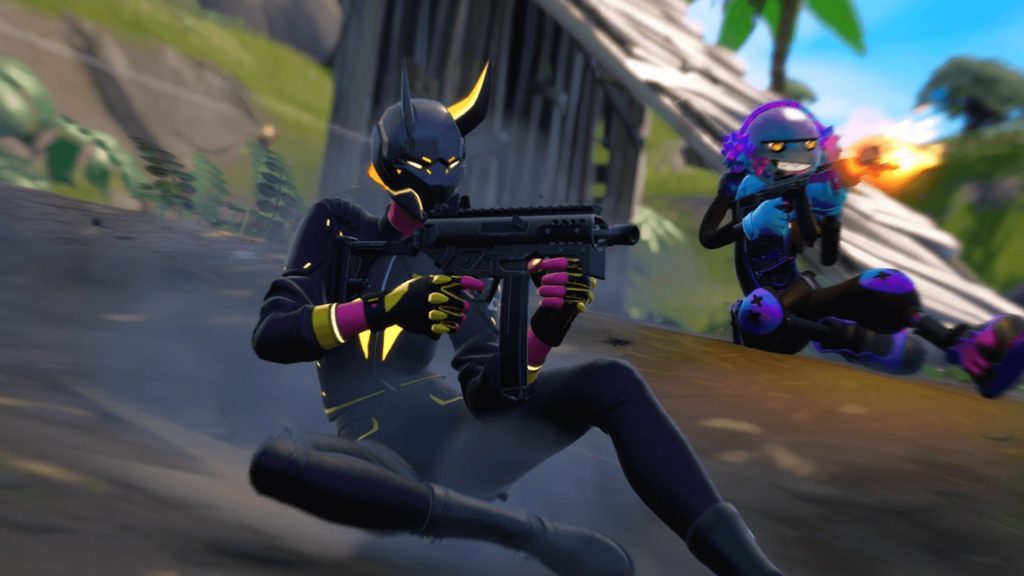 In this image we see two players in long shot in dark combat suits, sliding across the floor and shooting. We look at the characters from the side. The player in front has a black mask with horns on his head, and the one behind has a purple mask with a smiley face. The background has a motion blur and we see a green overgrown hill and two palm trees in the crop.