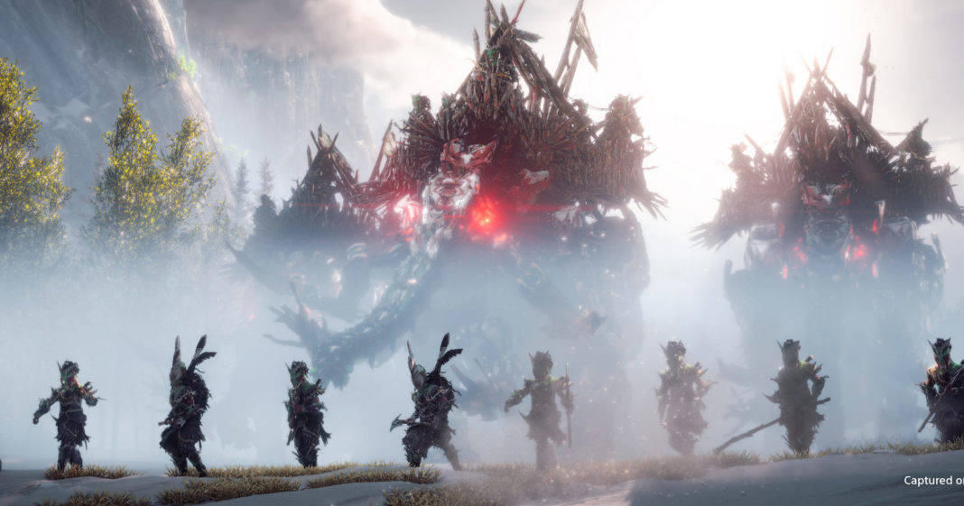 According to the latest rumors, a Horizon MMO is being planned. A co-op-style multiplayer might appear soon. In this screenshot, we see several characters standing in a row in front of giant creatures, ready to fight. They are shown in a long shot at the bottom of the screen, facing us, and have an Indian-like appearance with axes, sticks, and headdresses with large feathers coming out of them. Behind them, a strong mist can be seen and behind it, three staggered giant creatures with red glowing eyes that resemble elephants are shown. On top of the elephants are defensive structures where archers are stationed. On the left, green trees rise out of the fog, and in the distance, we can see a huge steep mountain range in the blur.