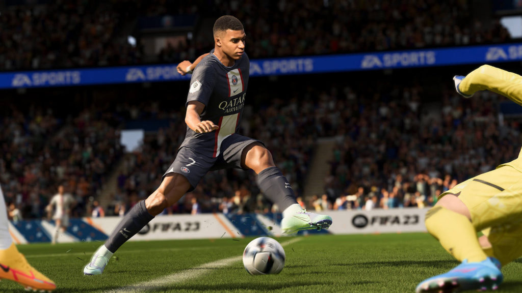 In FIFA 23 you'll experience breathtaking soccer matches together with your friends. This screenshot shows a southern soccer player in the center of the picture in the daylight with a blue, white, and red jersey in profile and short black hair, as he is about to kick the ball. Below him is green artificial turf with a white line. On the left, we see an orange shoe and on the right a goalward in a yellow outfit and blue shoes lunging for a jump. In the background, the audience in the stands, including advertising banners, is running horizontally through the image in a blur. The game is one of the best multiplayer PS5 games.