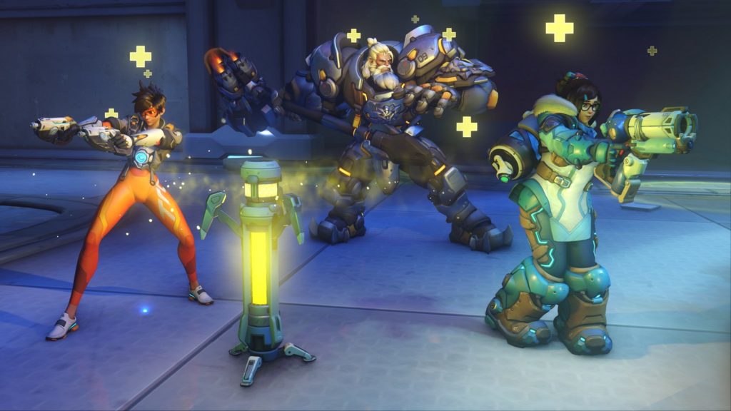 Here we see a screenshot from Overwatch 2 that shows three playable characters from the game in a long shot. All three players are looking in different directions. The left figure is very slim, dressed in orange with red sports glasses and holding two pistols to the front. The middle figure is arranged a bit further back in the picture and resembles a strong Viking with a blond beard and heavy battle armor. He has a large hammer in his hand. On the right, at the same level as the first figure, is a female figure with a green outfit and large green shoes. In her hands, she holds a rifle with a white barrel. In the foreground slightly to the left of the image, we see a yellow stick healing the characters so that yellow crosses pop up above their heads. They are in a metallic hall. The game can be played in multiplayer on the PS5.