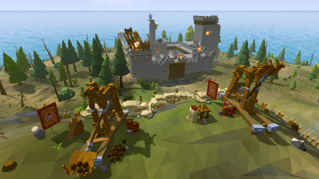 Build and destroy castles in Ylands, as seen here on the screenshot: In the foreground, two catapults are standing on a hill, just firing at a castle we can see in the background, which is already partially on fire. Behind the castle, the wide blue sea can be seen. Around the castle are numerous fir trees.