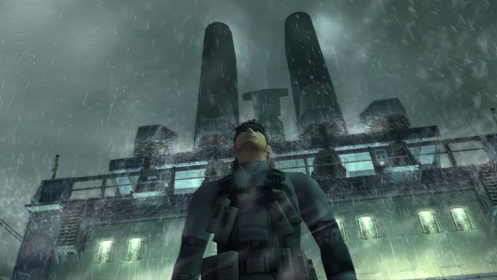 In this screenshot, we see the main character of Metal Gear Solid in the second part of the series from a strong bottom view and in the semi-close-up. He is shown centrally in the image as he stands in an industrial area. It's nighttime and raining heavily. His gaze falls forward. In the background, a factory building with illuminated windows and two huge chimneys rises into the dark sky.