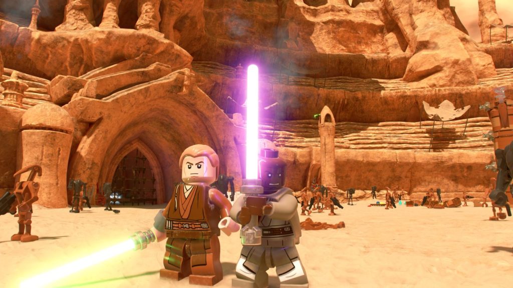 In this screenshot, we see two familiar Starwars characters in the center of the lower edge of the image in the wide shot in the foreground. The left character has brown clothes, blond hair, and a green lightsaber in his right hand. The right figure is a black character with light gray clothes. He holds a purple lightsaber with both hands. The two are standing on sandy ground in daylight, with numerous brown robot-like characters with black Assault Rifles depicted in the background. Further in the background, we glimpse a huge sand-colored mountain front taking up the entire frame and running vertically. On the left side, you can see a huge metallic gate through which you can enter the mountain.
