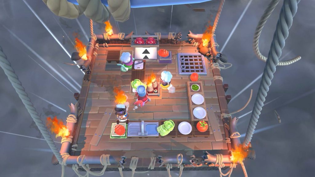 If you are looking for good entertainment and excitement at the same time with your friends, Overcooked! All You Can Eat should be one of the most suitable 2 player PS5 games for you. In this screenshot, we have a bird's eye view of the playing field, which consists of a square-shaped wooden kitchen floating in dark grey clouds. The kitchen hangs from four large ropes that rise out of the image to a blue hot air balloon visible in the crop. At the bottom of the kitchen, a long L-shaped kitchen counter is shown with several white plates, a wash basin, and various vegetables. The corners of the playing field are lit by four torches. At the top are more wooden tables and in the center are three different-looking players in the role of cooks, busily preparing the right menus in time.