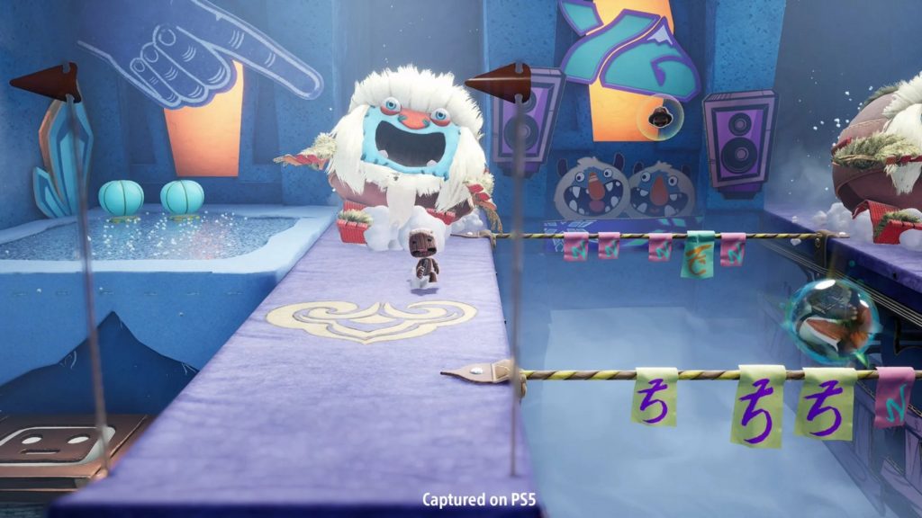 In this screenshot, the protagonist Sackboy can be seen in the center of the image in long shot. He is running away from a huge cute yeti-like white creature. The creature has white fur and its face has blue skin. With its mouth wide open, in which two round white teeth can be seen at the bottom, it is chasing the brown-colored Sackboy with its cute big sack-like head, which also has its mouth wide open. On a purple carpet leading toward the camera, the two run toward us. We are in a blue square-shaped room that resembles a toy world and has diverse playful different-colored motifs, such as a pointing hand or two face stickers that resemble the Yeti. To the left and right of the carpet, you can see the abyss, in which white mist.
