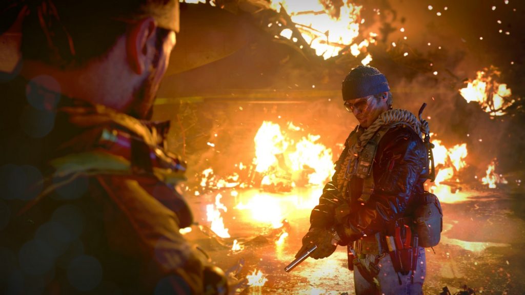 In this screenshot, we look over the protagonist's right shoulder at a soldier with war paint on his face, who is in focus on the right side in the image's middle ground. He is wearing blue jeans and a leather jacket that reflects numerous flames, which can be seen and in focus in the background of the image, taking up the entire frame in the back. The soldier holds a pistol with both hands, which he points to the ground. His gaze is also directed thoughtfully toward the ground, while his mouth is open. Apparently, the two soldiers are in the middle of a dialogue. We are in a house destroyed by fire in the evening. At the top of the image, a huge hole can be seen in the roof and we can see sparks of fire on the right side of the image. A gripping action spectacle and one of the best PS5 co-op games in 2022.