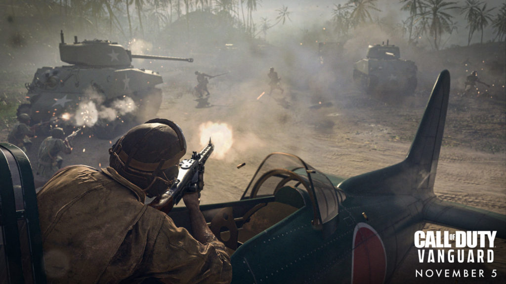 A screenshot from Call of Duty Vanguard can be seen. An intense combat battle is depicted in a dusty landscape during the day. In the foreground, we look over the shoulder of an Asian fighter pilot sitting in a blue airplane, which rises out of the picture in the foreground and can only be seen in the crop. He is firing an MG42 machine gun at onrushing enemy soldiers, who can be seen as dark silhouettes in the dust in the background. The soldiers are on a sandy road that curves to the right coming from the background and runs out of the picture to the right. On the left and right side of the picture, two M4 Sherman tanks can be seen in the wide shot. The one on the left is shown from behind and the one on the right is seen from the front. In the background are numerous palm trees, heavily shrouded in rising smoke and dust. On the bottom right, we look at the title of the game in white capital letters "Call of Duty Vanguard". Below that is the date in white capital letters "November 5".