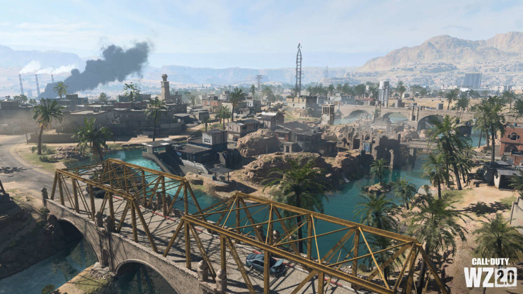 The new map of Al Mazrah is shown in this screenshot. We are looking at the large desert city from the top view. In the foreground, a large turquoise river is shown flowing from the lower left corner of the foreground to the upper right corner of the background. As it does so, it flows around an island in the center of the image a bit further in the background. Down in the foreground runs a large brown old stone bridge with a gold-colored metal fence on top of it, over which an asphalt road leads. Numerous different colored oriental-looking buildings and palm trees are in the city. At the top left of the image, there appears to be an industrial plant, as large black smoke rises from a chimney. Further back, slightly above the center of the image, a large radio antenna rises into the sky. In the distance, we can see yellowish mountains and above them the blue sky with some white clouds. It remains to be seen whether Warzone will also be released as Call of Duty for Nintendo Switch.