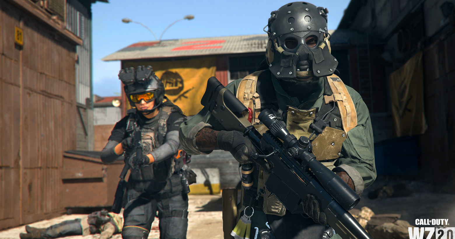 We look at a screenshot from CoD Warzone 2. The picture shows a soldier in an olive-green combat suit, a black full helmet in the semi-close-up a bit more on the right side of the picture during the day, as he runs straight towards the camera and looks into the distance in front of him. In his hands, he holds a metal sniper rifle. A bit further to the left, behind him, a female soldier in a bluish combat suit and a black helmet with an orange visor is running toward us. She holds an Assault Rifle in her hands. Both characters move past dead soldiers lying on sandy ground. On the left edge of the picture, the large brown locked iron gate of a factory building looms into view. Also in the background is a similar-looking hall with a light gray corrugated iron roof, on which orange graffiti has been sprayed. On the facade, there is a huge orange fabric banner with a logo showing a Kalashnikov. We also find such a banner on the right side, which is attached to a very dark appearing building facade lying in the shade. Within 10 years, Call of Duty for Nintendo Switch is scheduled to be released.