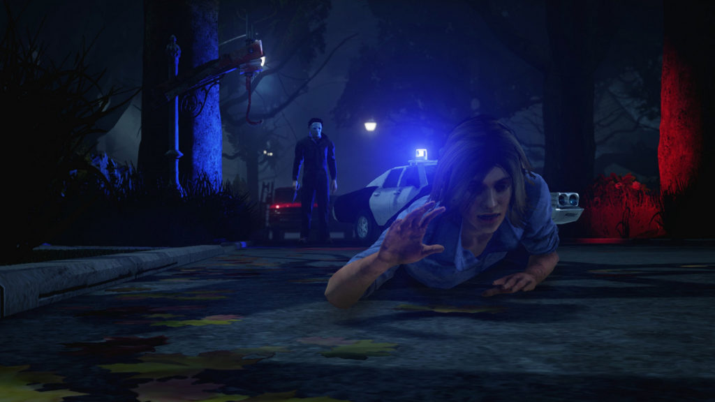 In this screenshot from Dead by Daylight, a female playable character can be seen in the foreground of the image from the front in a long shot, crawling along a street in our direction. The person is wearing a light blue blouse and has shoulder-length blonde hair. The scene takes place in a section of forest in the autumn season at night. Leaves are lying on the road. In the background stands another playable character in the guise of Michael Myers with his iconic white mask and a large kitchen knife, ready to kill his victim. To the right of him is an American police car with blue glowing lights. It casts a red spotlight on a tree on the right side of the image. We can also make out the black silhouettes of individual tree trunks on the left edge of the picture.