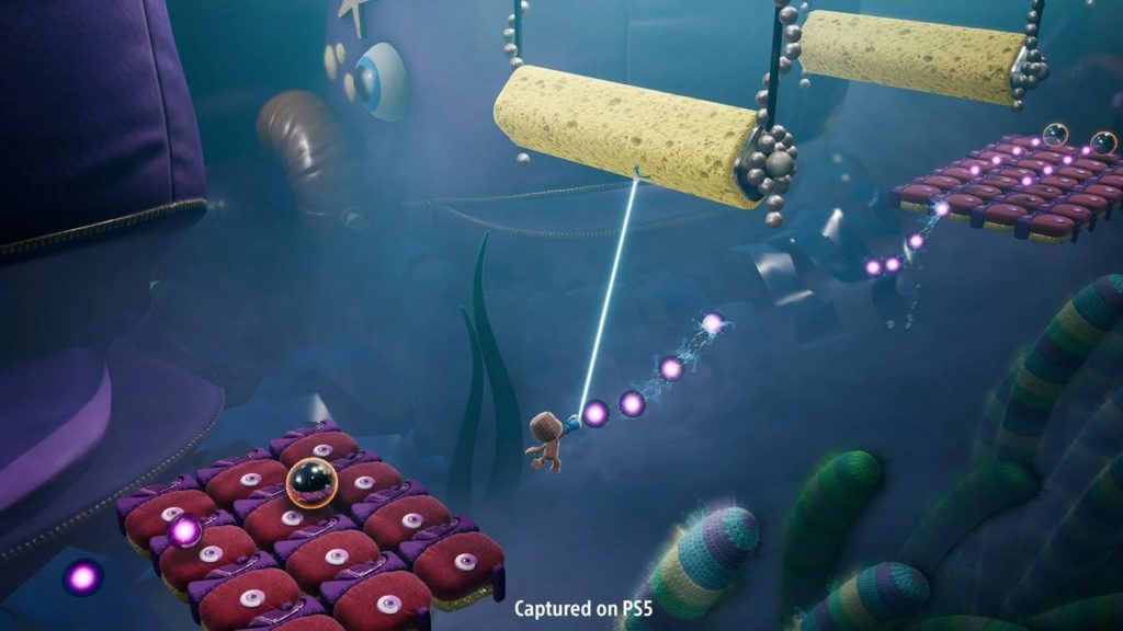 You see the brown textile hero Sackboy with his big cute head diagonally from behind in the lower right center of the picture in the long shot. He is swinging with the help of a grappling hook through a dark blue, gloomy-looking world that consists of various toy-like elements. The world is completely three-dimensional with very pretty graphics, but the game level runs linearly from the lower left front to the upper right back here. Between the player are platforms made up of red and purple colored cushions. Sackboy hangs on a kind of rope, at the end of which there is a hook attached to a kind of yellow sponge roller. Now he has to jump to a second roller with enough momentum to finally get to the other platform. This title can become one of the games like It Takes Two with up to four players.