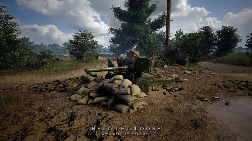 In the center of this screenshot, a green AT-Cannon is shown in total view behind a sandbag barricade facing the left side of the image. It stands on brown sandy ground, which takes up most of the image. To the right of the cannon, numerous spent shell casings lie on the ground. Also in this picture, there is bright sunshine and a few white clouds appear in the blue sky. A puddle of water is shown in the lower right foreground of the picture. Further back in the image a green landscape with numerous different green trees and bushes can be seen and behind it, there is a field. To the right, a sandy path leads out of the background right to the front of the image. Further, in the distance, a horizontally running stone wall can be seen and behind it an avenue of trees.