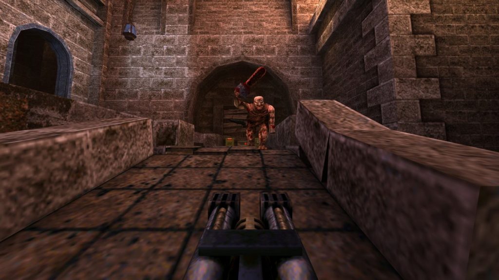 The player is shown here in first-person view. He holds a futuristic submachine gun with two barrels in his hand, which we can see in the center at the bottom of the screen. The player is standing on a stone bridge, which is located in a medieval castle courtyard and is illuminated by the evening sun. In front of him, at some distance, stands a bloody naked beige monster with a bald head and distorted face, holding a bloody chainsaw in the air with his right hand. On the left side, we can see a gate with a bridge leading to it, and behind the monster, there is also a barricaded entrance under a round archway. The game Quake is available as a remaster for the PlayStation 5 and is one of the highly recommended PS5 split-screen games.