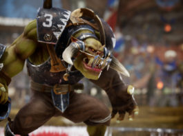 We can see an Orc from Blood Bowl 3 in this screenshot. He is shown in the semi-close-up in the half-profile on the left of the image. The muscular, green-skinned creature wears brown cloth pants and has football-like iron armor. In front of his mouth, the orc wears a kind of protection in the form of a brace. In his right hand, he holds a football brown-leather ball device. With a pouty mouth and sharp teeth, he looks to the right. In the background, the tribune is shown with blue banners in a solid blur. The scene takes place during the day. A release date is confirmed for February, and there will likely be crossplay.