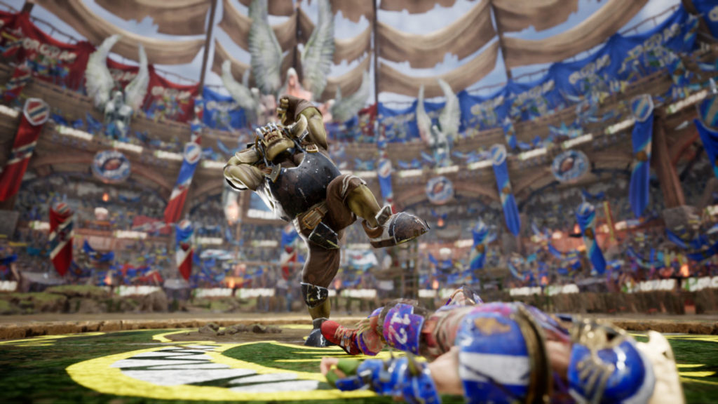 From a strong bottom view, we see a powerful orc with a similar appearance to the one described in an earlier screenshot from BB3. He stands in the middle of the playing field during the day and is in front of a blue human player lying on the ground, which we can see out of focus in the foreground on the right. The orc player is about to strike with a huge axe. In the background, the cheering crowd in the stands with numerous blue and red banners can be seen out of focus. The stadium has a very round shape.