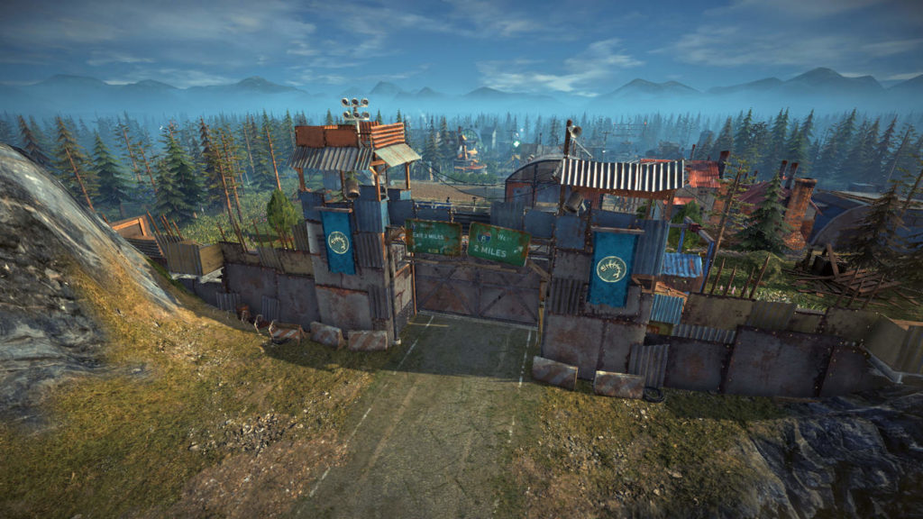 Play Surviving the Aftermath and experience the dangers of the post-apocalypse, as shown in this screenshot: From an elevated position we look at a heavily barricaded entrance to a city. We see a road coming from below leading to a large rusty gate with two large observation towers to the left and right. Rusty high walls extend from the towers and continue around the city. Behind the walls in the background, several different colored roofs of houses can be seen together with many fir trees rising into the sky. In the foreground of the picture, there are steep cliffs and in the distance, we can see a mountain range passing through the picture, which lies in the light blue mist. Above it, we can see the blue sky with clouds.