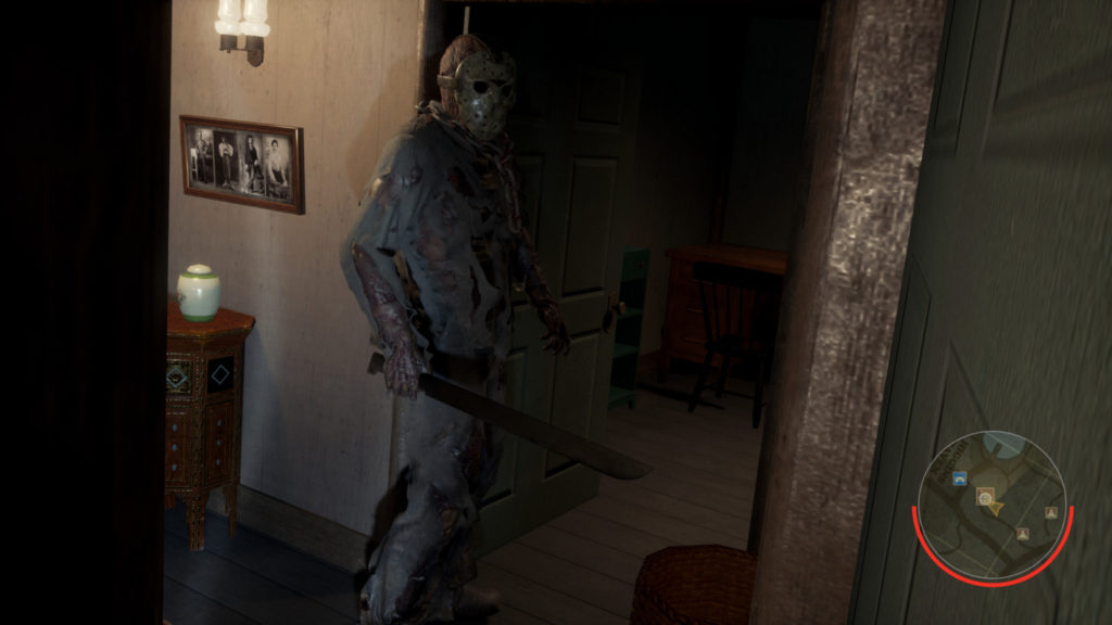 A screenshot from Friday The 13th The Game is shown: We see the well-known killer Jason Vorhees with his iconic field hockey mask. He is shown on the left of the image in a long shot, standing at an open green door in an apartment. His gaze falls to the right. In his right hand, he holds a long straight sword and he wears a bloodstained gray jumpsuit. The apartment is very comfortably furnished and consists of a plank floor and white wallpaper. On the left edge of the picture, there is a wooden commode with a white vase and above it a picture and a sconce. In the background of the picture in the other room, there is a wooden desk with a black chair and to the left of it a green chest of drawers. On the right edge of the picture, another green door can be seen in the crop.