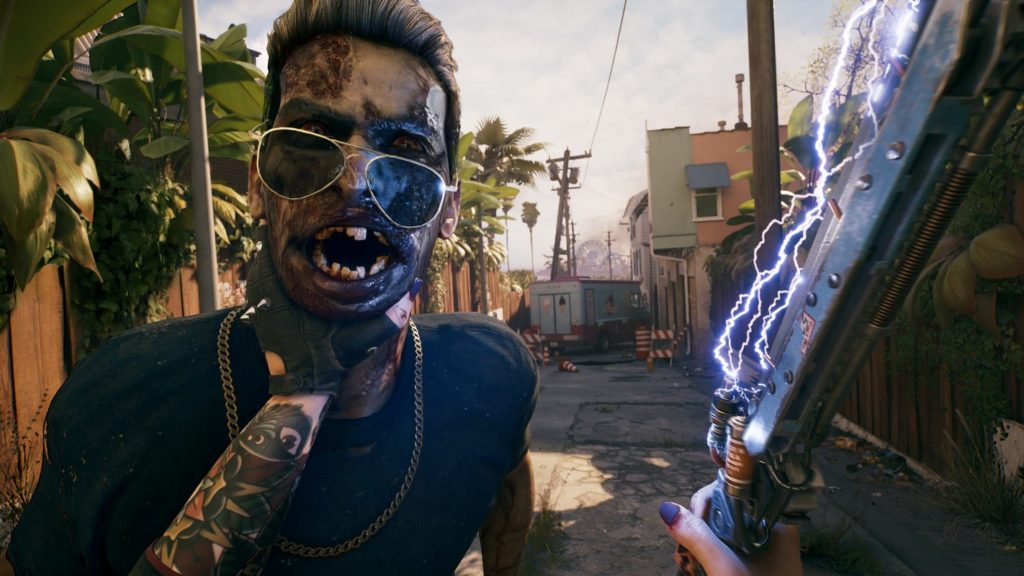 We are the player in first-person perspective in a side alley in Los Angeles. With our left hand, we are strangling a male zombie with a blue T-shirt, black hair, and aviator goggles. With his mouth wide open, we catch sight of his virus-ridden teeth and decayed skin, and red eyes. Our character has numerous colorful tattoos on his left arm and purple nail polish on his right hand indicates that we are playing a female character. In our right hand, we hold a blue flashing self-crafted metal blade. To the left and right are high wooden fences that run along the alley into the background. There we see an abandoned ambulance and different colored house facades on the right. The sun is shining and gives the scene a summery mood. This zombie survival shooter offers you extremely intense and brutal gameplay.