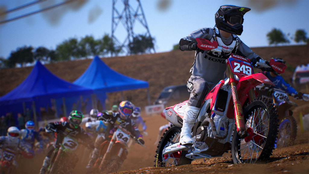 In this screenshot from MXGP 2021, we look up from a brown muddy race track in the middle of the day to a column of colorful racers in helmets just turning a corner and passing us on colorful motocross bikes. All the riders are blurry in this picture, whereas the number one on the right side of the picture is in sharp focus in a long shot. He has a gray jersey, a black helmet, and a red-colored motorcycle, and mud is spraying toward us, causing dirty spots to appear on the camera lens in the blur at the top and bottom of the image.