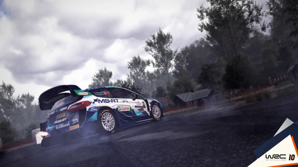 Experience one of the best racing games for PS5 with this rally simulation. In this screenshot, we can see a blue-white race car from a bottom perspective with numerous different colored sponsor logos on it. The car has just passed us and is turning right around a bend, so we can see it in half-profile from behind. The road is asphalt and gray. In the background, we can see several brown wooden houses with sloping roofs in the center of the picture and around them a forest with numerous tall green trees. In the bottom right corner of the picture, we can see the logo of the game "WSC 10" on a white triangle.