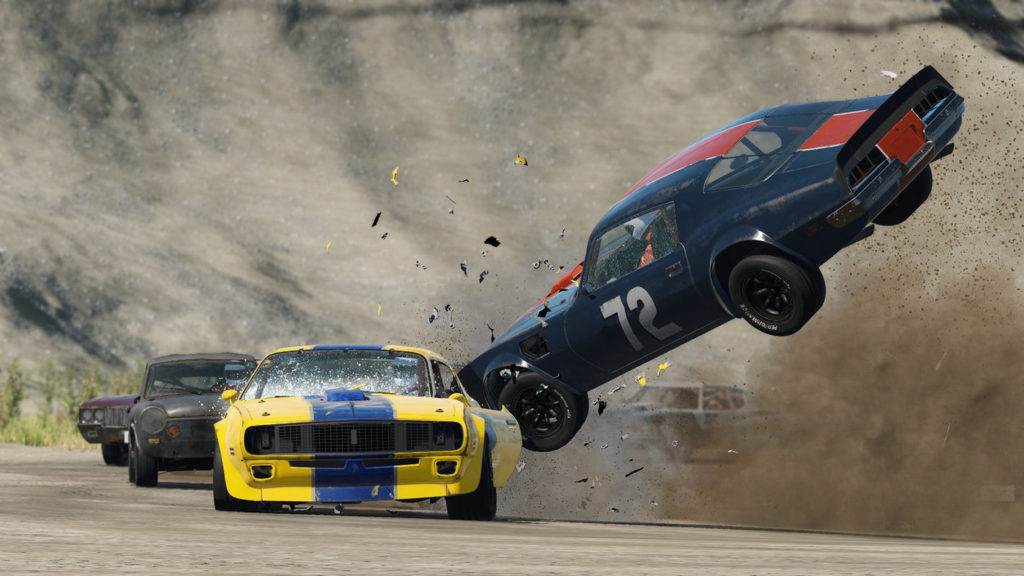 PS5 racing games like Wreckfest focus less on proper driving and more on destruction and chaos. In this screenshot, we see a column of racing cars on a brown racetrack during the day. The cars are driving around a curve. We look from the front at the first two cars. There, a yellow-blue car, which we see on the left side of the screen, is just rammed by a dark-blue-red car, so that numerous splinters of both cars fly into the air and also the windshield of the yellow car is broken. The right car also flies into the air due to the crash, which causes quite a lot of dirt and dust to be stirred up. In the background, we see a mountain range in the blur, which covers the entire frame.