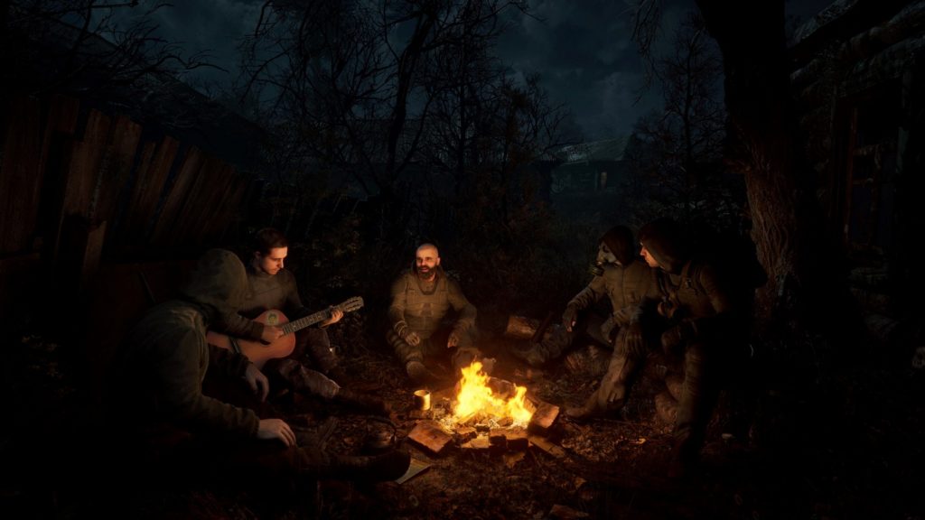 In this screenshot, we see five men in dark uniforms in a long shot sitting around a campfire in the middle of the night. One of the men on the left is playing an acoustic guitar, while the man with a bald head and beard sitting in the middle is telling a story and looking in our direction. The person to his right is wearing a gas mask. Around the men, the scene sings in darkness.