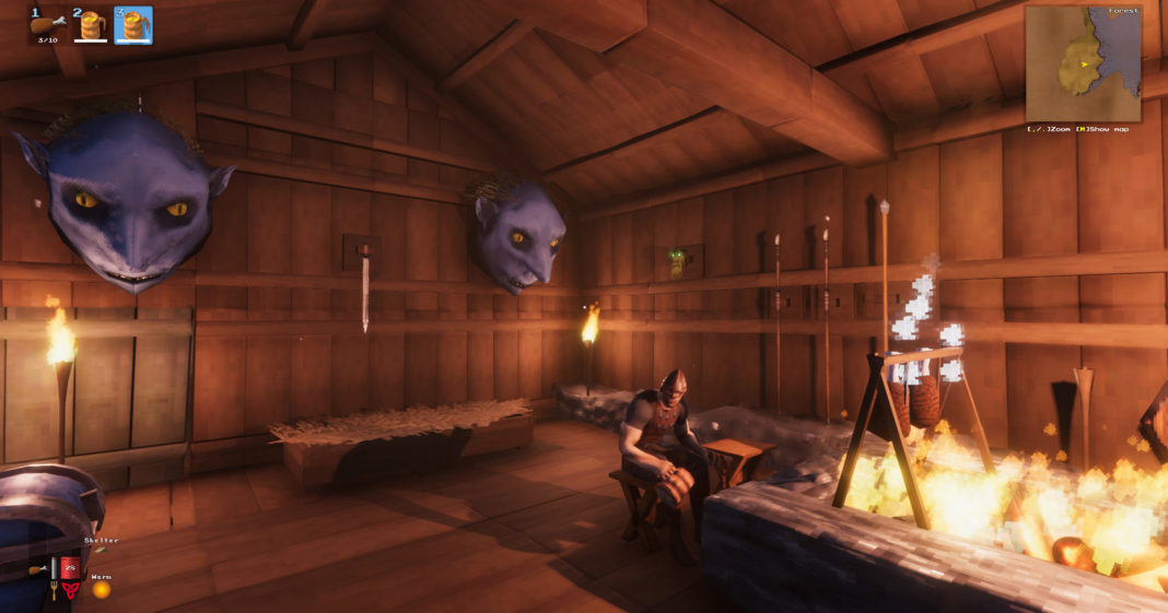 In this screenshot, we are inside a Viking longhouse built entirely of wooden planks. We are standing in the corner of the room as the viewer. On the far wall are two large blue troll heads as trophies. Next to them are two burning torches. In the lower-left corner, a blue iron chest can be seen in the cut. The building has a corner roof, which we can see at the top of the picture. Our gaze wanders to the center of the picture, where a Viking in brown leather armor and a pointed iron hat is sitting on a wooden chair. He looks at us as the viewer. Directly in front of him is a huge rectangular fireplace, which can be seen in the lower right of the picture in the crop. In the fire, a holding device is attached, to which two large pieces of meat hang down getting grilled. White pixelated smoke is rising. There are already many Valheim biomes. Now the game will soon be expanded with another region, the Ashlands. The title will also be available for Xbox early this year and will support crossplay.