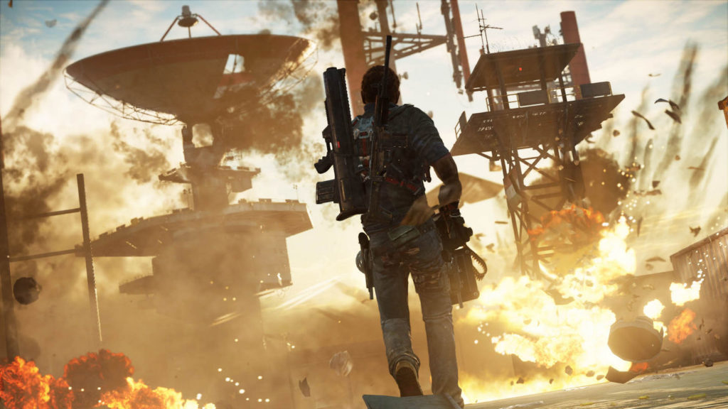 In this screenshot from Just Cause 3, we see the protagonist in the third person in the center of the image in a long shot. He is running with a minigun in his hands towards a military base during the day, which he is shooting at the same time. He has short black hair, and wears a blue shirt and blue jeans. On his back, he has an Assault Rifle and a Grenade Launcher. On the left in the background, a huge radar station with a satellite dish rises into the air, and to the right of it, we see a surveillance tower, at the base of which a violent explosion is taking place. Also at the bottom left of the image, we glimpse an explosion in the crop, and throughout the base in the background smoke projectiles shoot through the air and the scene is enveloped in fire and smoke. One of the most insane sandbox games on Steam.