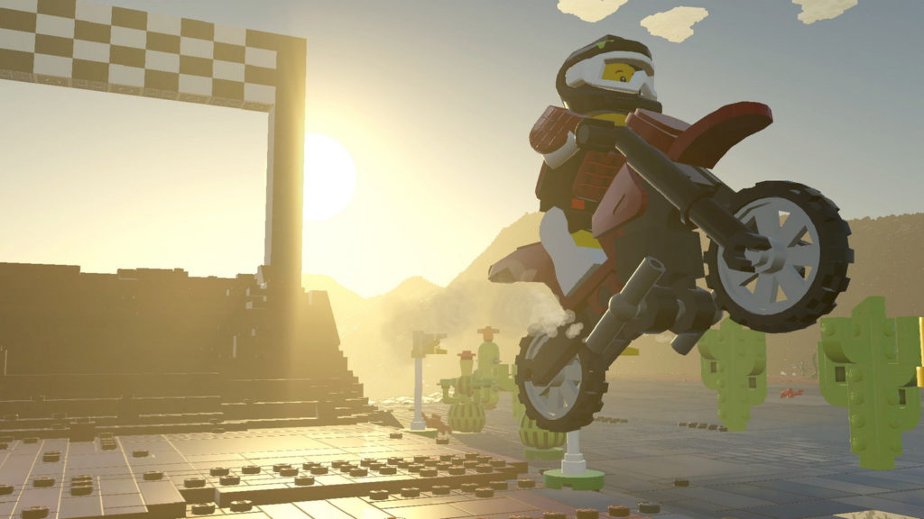 Build your brick world the way you like it. This screenshot from Lego World shows a player wearing a red jersey, white pants, and a white and black helmet on a motocrosser on the right side of the screen in a long shot. He's just jumping with his motorcycle to the front right, which gives us a half-profile view of him from the right side. On the left side of the image is a built race track made of Lego bricks, which leads from the back left to us in the foreground. In the background, the blue sky is shown and the setting sun shines directly into our eyes, creating a very nice evening atmosphere. At the right back, we can see a mountain range and numerous placed light green cacti to the right of the player.
