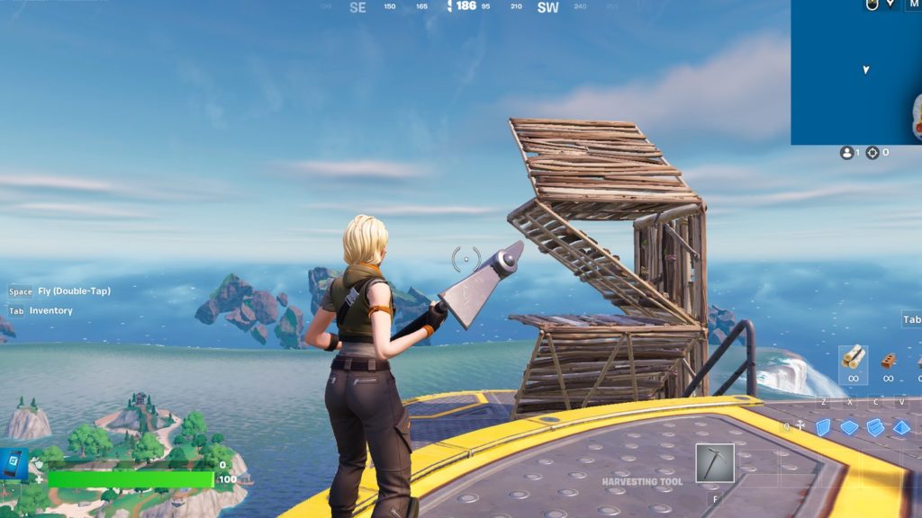 The player is on a colorful bright island during the day and is standing on a round metal platform with a yellow border. He is shown in the third-person centered at the bottom of the image and is a female character with blond hair and brown combat outfit and a pickaxe. The blue sky can be seen in the background. The character is looking at a wooden construction he has just erected. This is a 90s build. The player has built several ramps on top of each other, always turned to the right at a 90-degree angle, in order to reach the top vertically. In doing so, he has built walls around the ramps to protect himself from enemy gunfire. Fortnite building tips also include this building technique.