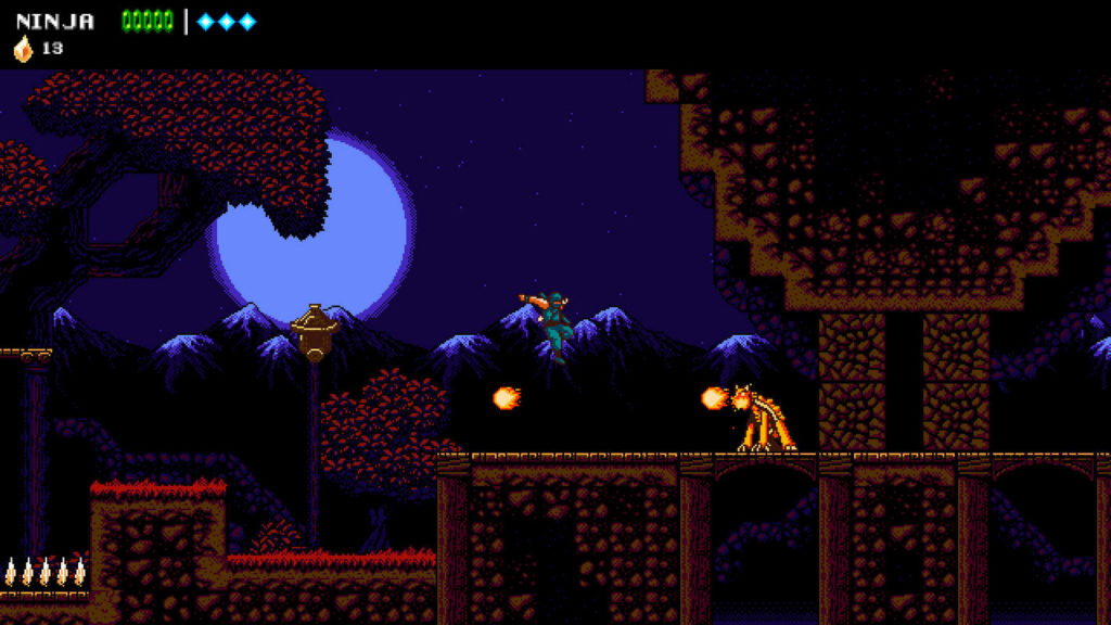 This screenshot shows the player as a ninja in turquoise battle gear in the wide shot in the center of the image. He is in a pixelated diabolical-looking red level that takes place in the middle of the night. In the background, there are pointed dark mountains shone by the moon, above which the large white-bluish moon is shown in the upper left corner. The ninja is currently jumping over fireballs spat out by a demonic red and yellow turtle-like creature. The playable foreground of the level features rectangular earthy structures. On the left is a tall streetlight, and in the bottom left corner we can see black spikes on the ground.